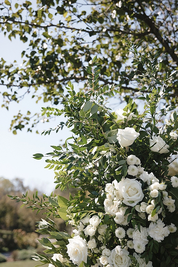 Wedding floral arrangement of white rose wedding flowers with lush greenery, set outdoors against tree branches and blue sky
