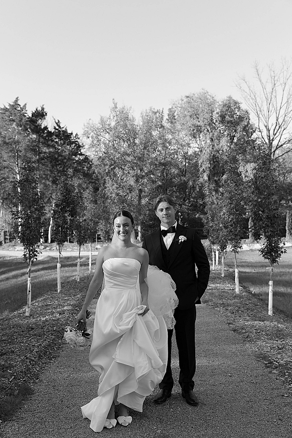 Couple portrait in black and white, bride in strapless dress and veil holding bouquet beside groom in tux on a tree-lined path
