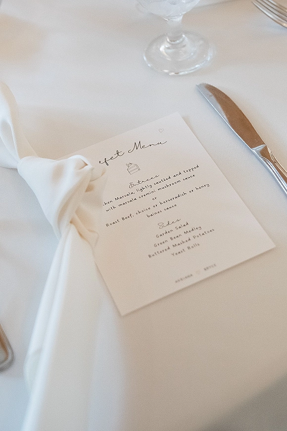 Wedding menu card with calligraphy text on a white napkin beside a knife and wine glass on a white tablecloth place setting