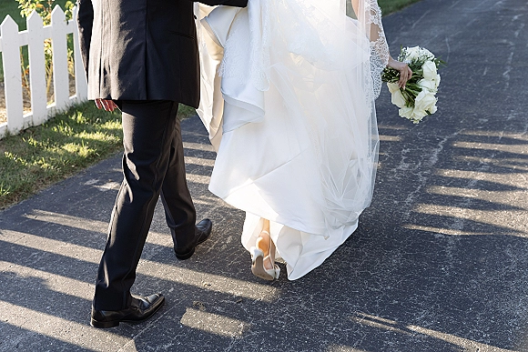 Wedding couple walking hand in hand, bride and groom walking away with white rose bouquet and veil along a sunlit path by a picket fence
