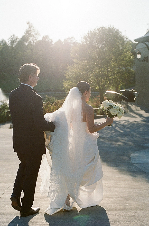 Newlywed walk as bride and groom stroll away hand in hand, veil and bouquet glowing in sun flare on a tree-lined lakeside path