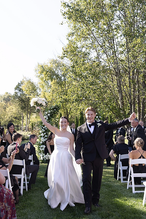 Wedding recessional of bride and groom walking aisle, bride lifting bouquet and groom raising arm, guests on white chairs by floral arch outdoors