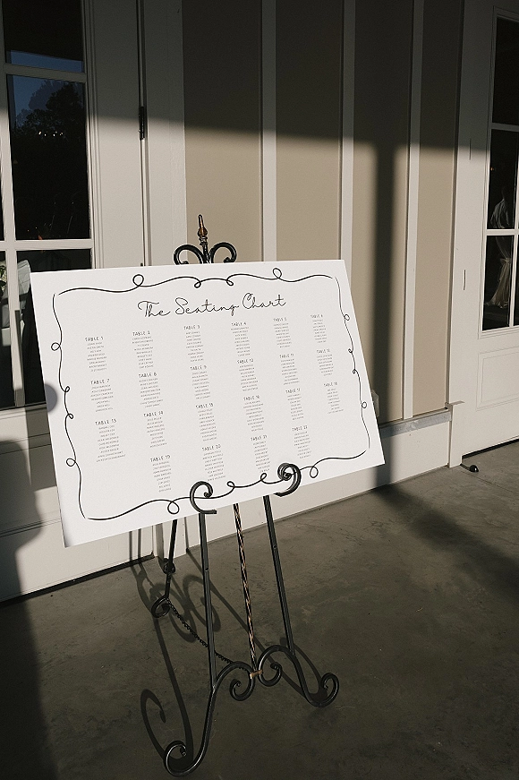 Wedding seating chart with calligraphy seating chart lettering on a white board, displayed on a wrought iron easel by glass doors in sunlight