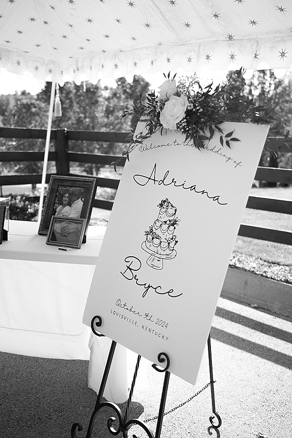 Wedding welcome sign with calligraphy on an easel, topped with roses and greenery, beside framed photos under a canopy tent outdoors