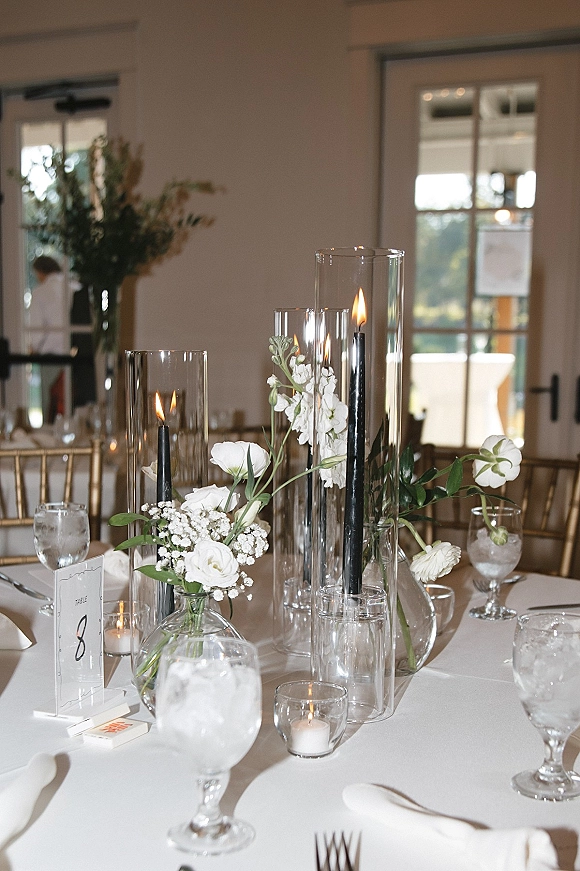 Reception tablescape with a wedding table centerpiece of white florals, bud vases, and black taper candles glowing by window-lit glass doors