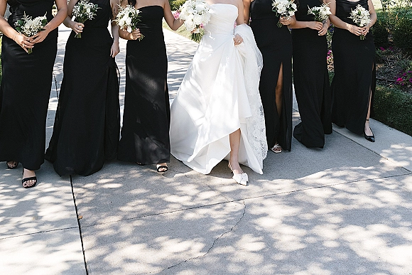 Bridesmaids photo with bride in strapless wedding dress and veil, flanked by black bridesmaid dresses holding white rose bouquets on a tree-lined walkway