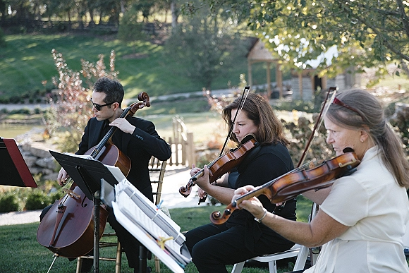Wedding ceremony musicians in black play violins and cello from sheet music on stands on an outdoor lawn by a gazebo and trees