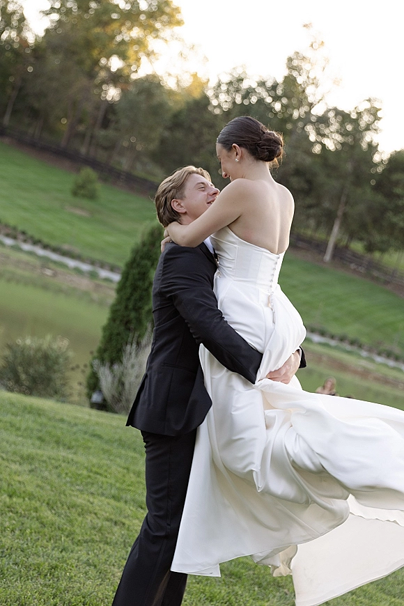 Couple portrait of groom lifting bride in a strapless wedding dress, her train flowing on a grassy hillside near a pond and trees