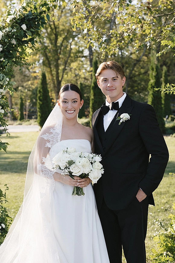 Couple portrait of bride in strapless gown with lace veil holding white rose bouquet, smiling beside groom in black tuxedo in a garden