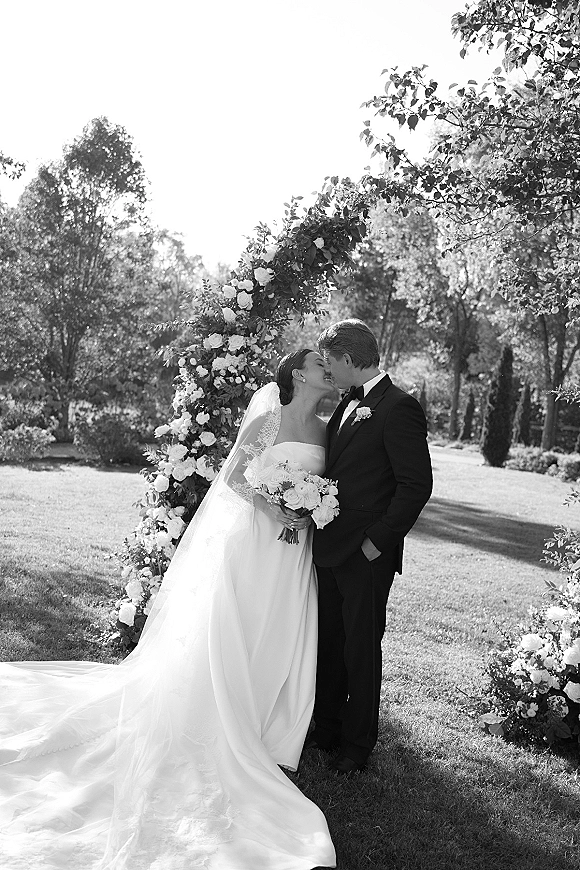 Wedding kiss portrait of bride and groom kissing beneath a rose floral arch, bride in strapless dress with long veil holding bouquet on garden lawn