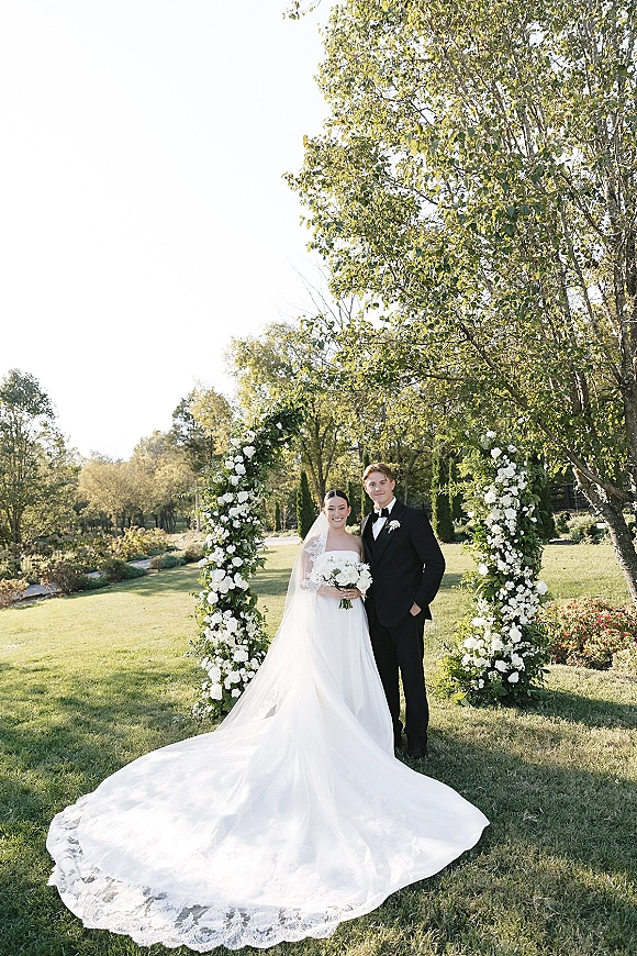 Couple portrait of bride and groom standing under a wedding floral arch, her lace veil and long train in sunlight on a garden lawn