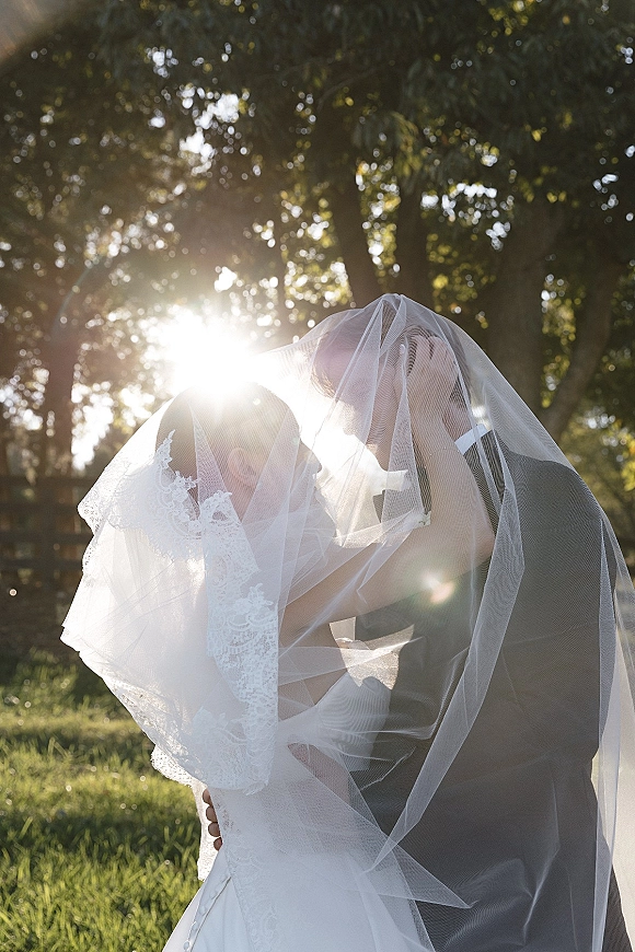 Wedding kiss portrait of bride and groom under a bridal veil, lace dress and suit, backlit by sun flare with trees and grass behind