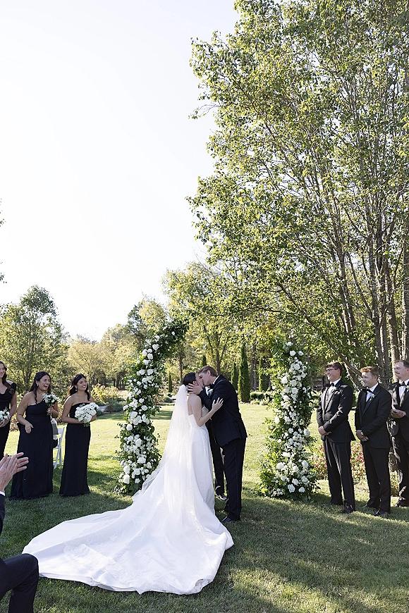 Wedding kiss as the bride and groom kiss under a white rose and greenery ceremony arch, veil flowing on a sunny garden lawn