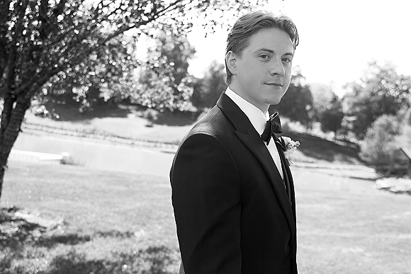 Groom portrait in a black tuxedo with bow tie and white boutonniere, standing on a sunlit lawn with trees and hillside behind