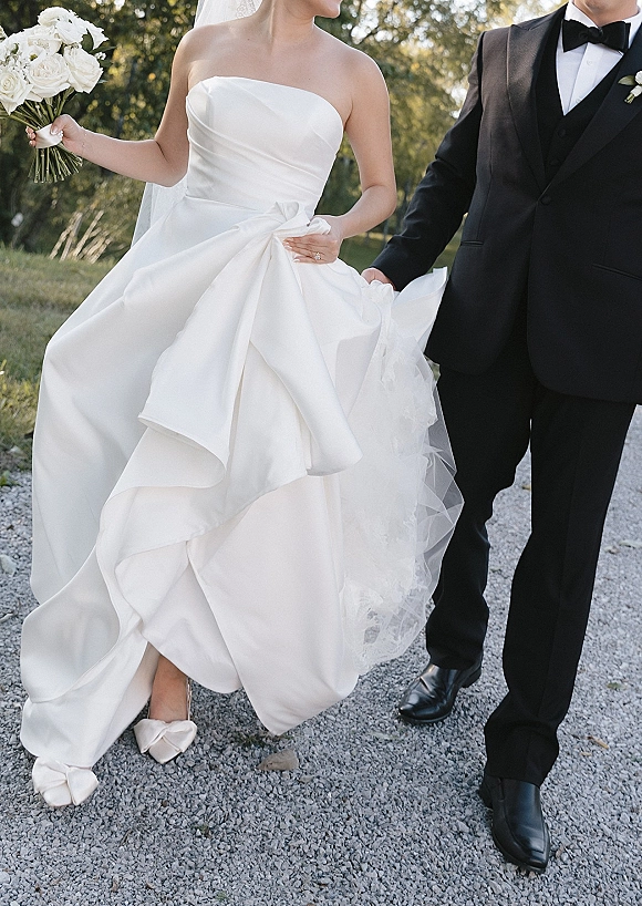 Couple portrait of bride and groom walking, bride holding a white rose bouquet with veil trailing on a tree-lined gravel path