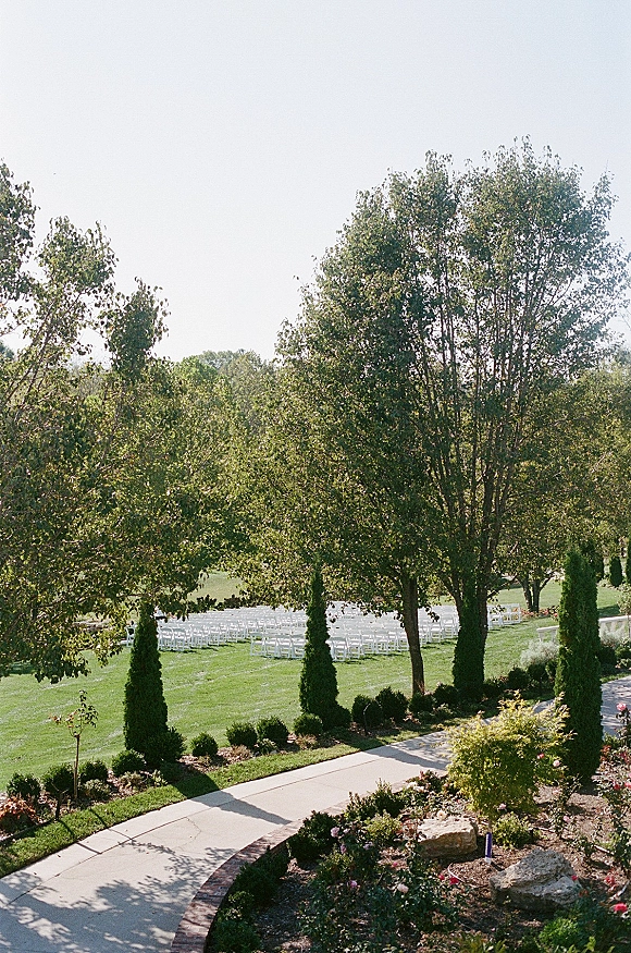 Outdoor ceremony setup with rows of white ceremony chairs arranged on a green lawn beside a curved walkway and garden landscaping