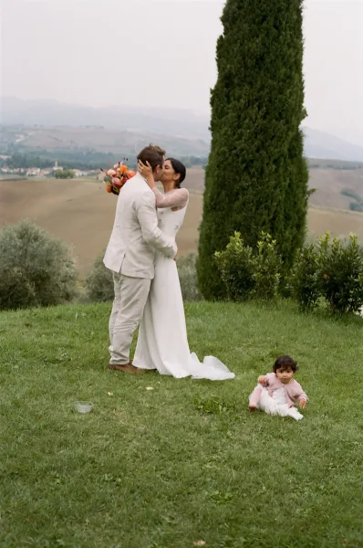 Wedding kiss outdoors as bride in a long sleeve gown holds a colorful bouquet while groom in beige suit cradles a toddler on a grassy hilltop
