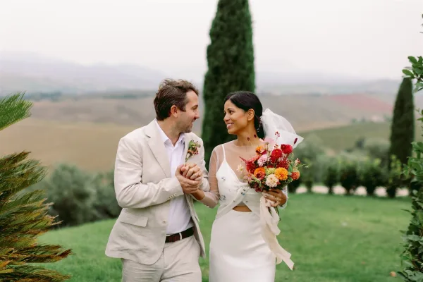 Couple portrait of bride and groom holding hands, her wildflower bouquet and veil flowing as they walk on a lawn with hills and cypress tree behind