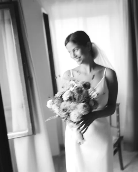 Bridal portrait in black and white of a bride in veil holding a mixed flower bouquet, lit by window light beside a framed mirror indoors