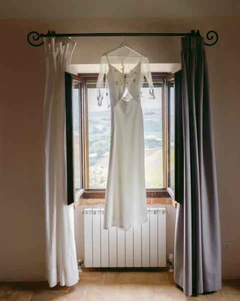 Wedding dress hanging photo of a bridal gown on hanger with embroidered bodice and sheer sleeves by an open window with countryside view