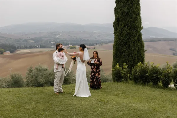 Ceremony moment at an outdoor wedding ceremony as bride touches groom’s face while he holds a child on a grassy lawn with mountains behind