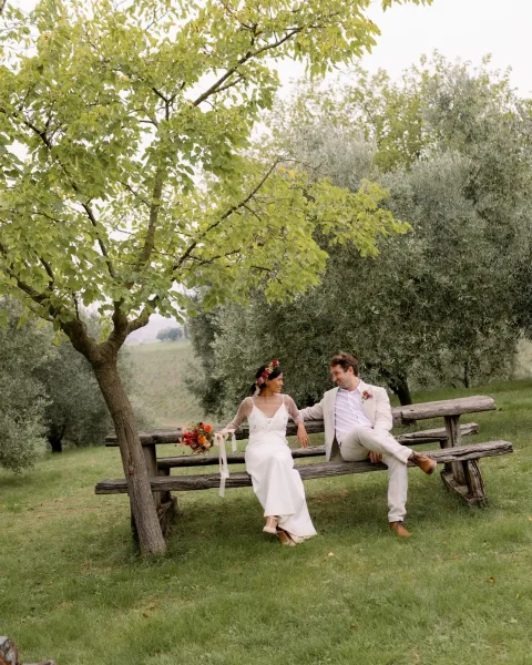 Couple portrait of bride and groom sitting on a rustic wooden bench, bride holding bouquet in an olive grove with rolling hills