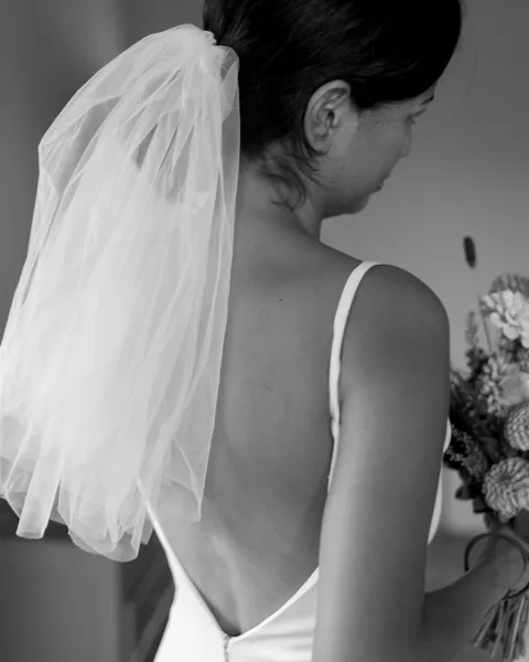 Bridal portrait of a back view bride in a low back spaghetti-strap gown with short veil and bouquet, standing by an indoor doorframe