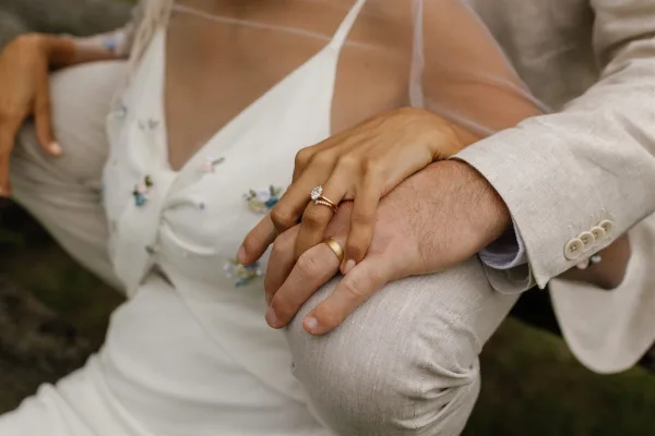 Wedding rings close-up showing an oval diamond engagement ring and gold band on bride’s hand resting on groom’s linen jacket, blurred greenery behind