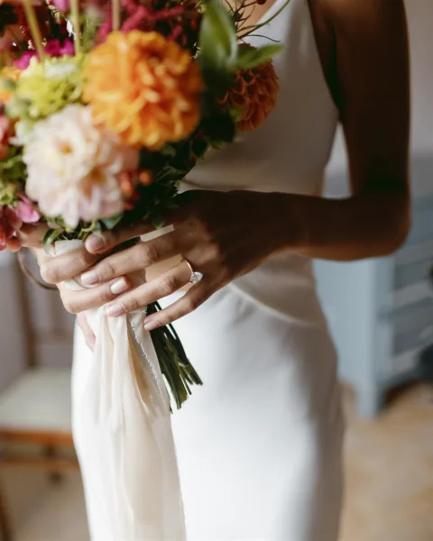 Bridal bouquet with colorful wedding bouquet of pink and orange blooms, ribbon wrap and ring detail in soft indoor light