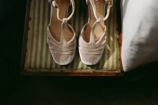 Bridal shoes with wedding heel details, velvet ankle-strap sandals resting on a striped chair cushion beside white fabric