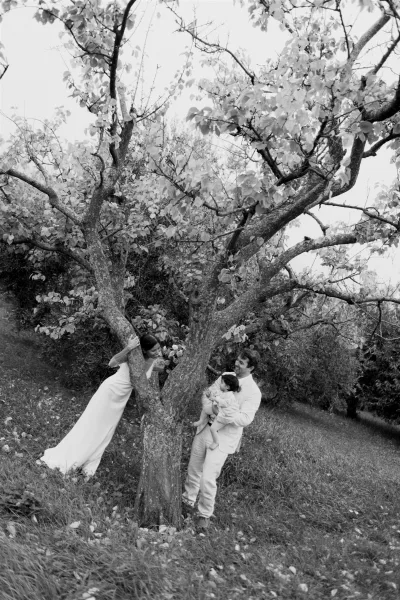 Family wedding portrait in black and white of bride and groom with baby by an orchard tree, bride holding bouquet, groom in suit with boutonniere