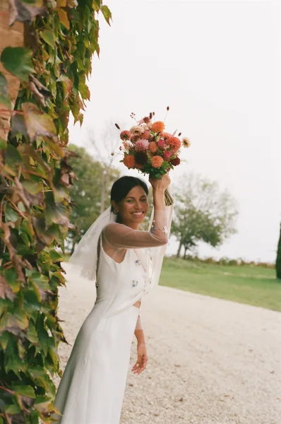 Bridal portrait of a smiling bride holding bouquet overhead, her veil and white dress set against an ivy wall outdoors