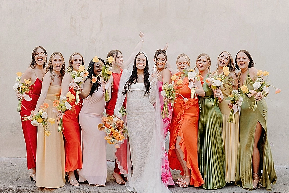 Bridesmaid group photo with bride and bridesmaids in colorful bridesmaid dresses, holding bouquets with ribbons against a stucco wall backdrop