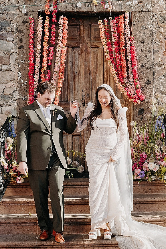 Wedding recessional of bride and groom walking hand in hand down stone steps, her cathedral veil and lace gown framed by floral garlands