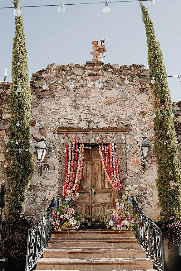 Ceremony entrance decor with floral garlands draped over wooden double doors, string lights above, and arrangements on stone steps by cypress trees