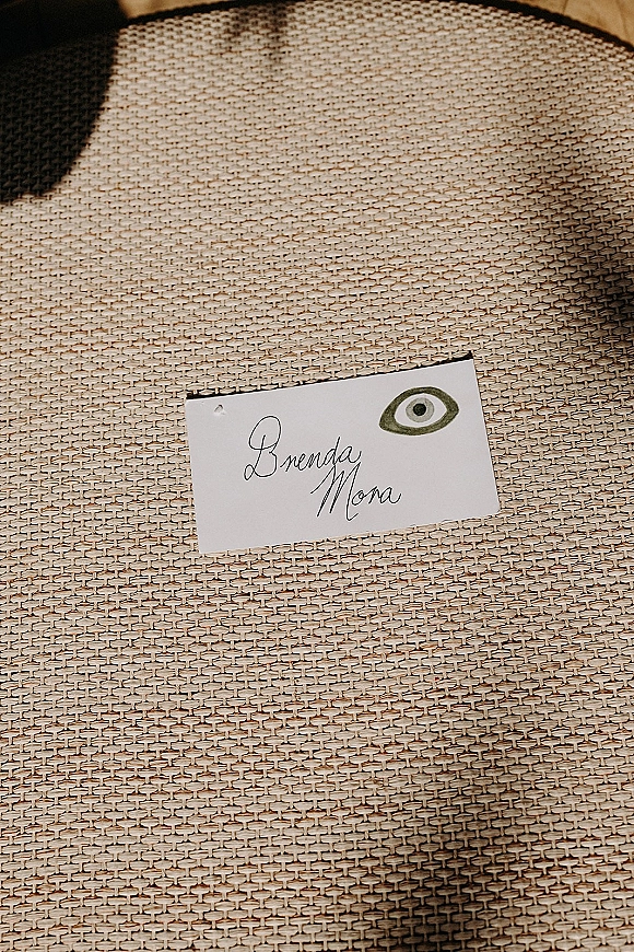 Wedding place card with calligraphy place cards styling, featuring an eye illustration resting on a woven chair seat in soft shadow light