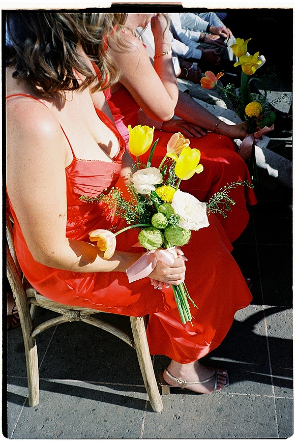 Bridesmaid portrait in a red bridesmaid dress, holding a tulip bouquet with greenery and a pink ribbon, seated among wedding guests