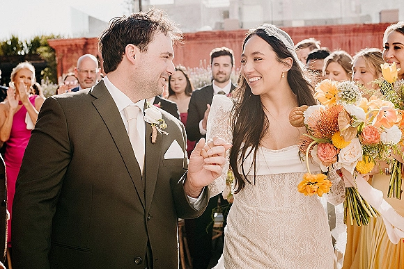 Wedding recessional as bride and groom walk hand in hand past cheering guests in a sunny brick courtyard, bride holding bouquet