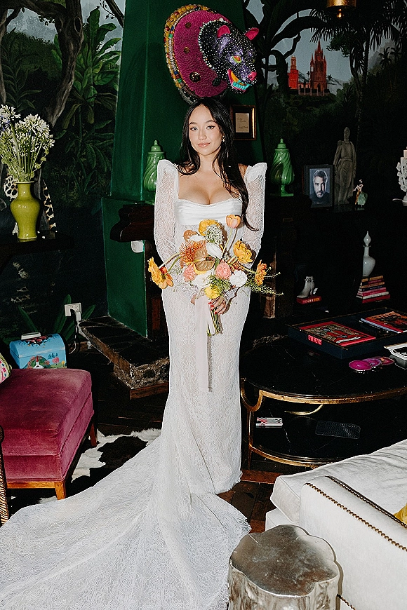 Bridal portrait of a bride holding bouquet in a long sleeve lace gown with square neckline, posed by a dark green mural wall and fireplace indoors