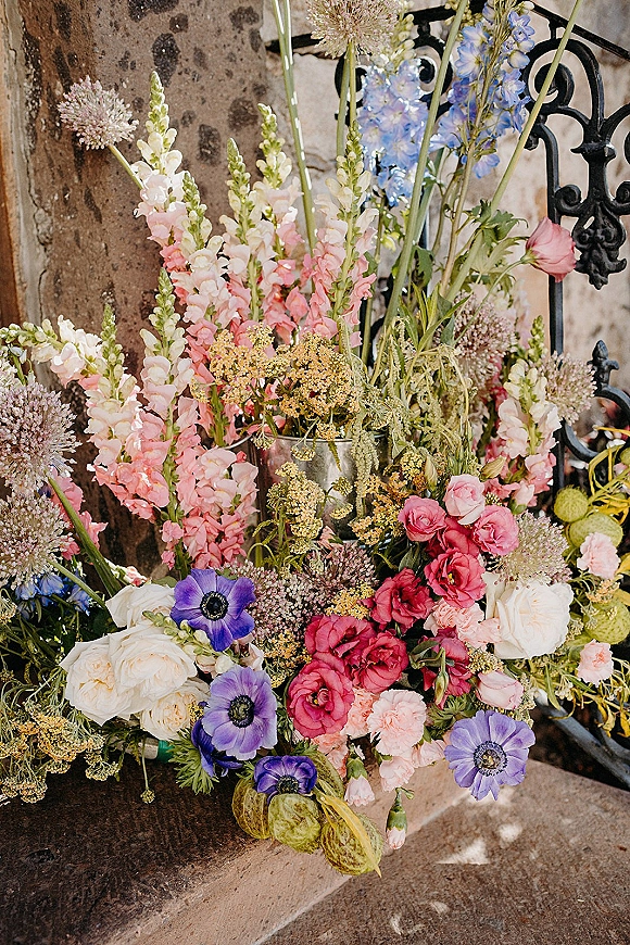 Wedding flowers arranged in a metal vase, with pink snapdragons, white roses, purple anemones, and delphinium by a stone wall