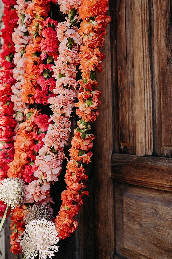 Wedding floral installation with hanging flower garlands in coral, pink, and orange cascading over a rustic wooden door backdrop