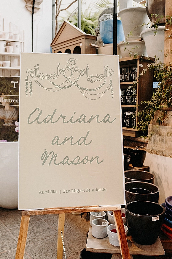 Wedding welcome sign with calligraphy welcome sign lettering and floral illustration on a wood easel in a plant shop with potted greenery