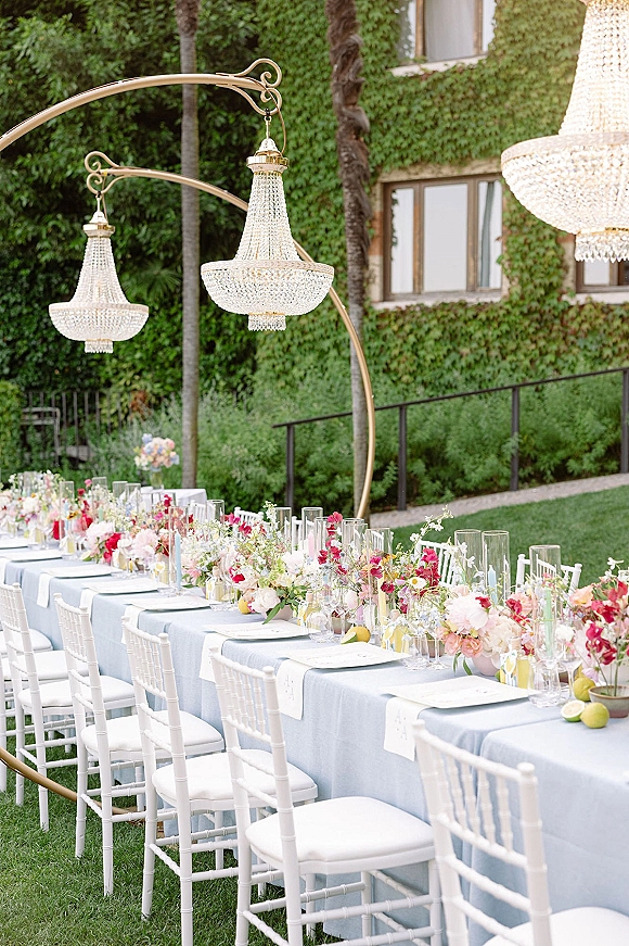 Reception tablescape with a long banquet table on a garden lawn, light blue cloth, pastel flowers, hurricane vases, crystal chandeliers, and citrus accents