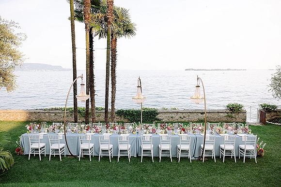 Reception tablescape with long banquet table setup on a lawn, light blue linens, lemons and florals, chandeliers glowing against an ocean view