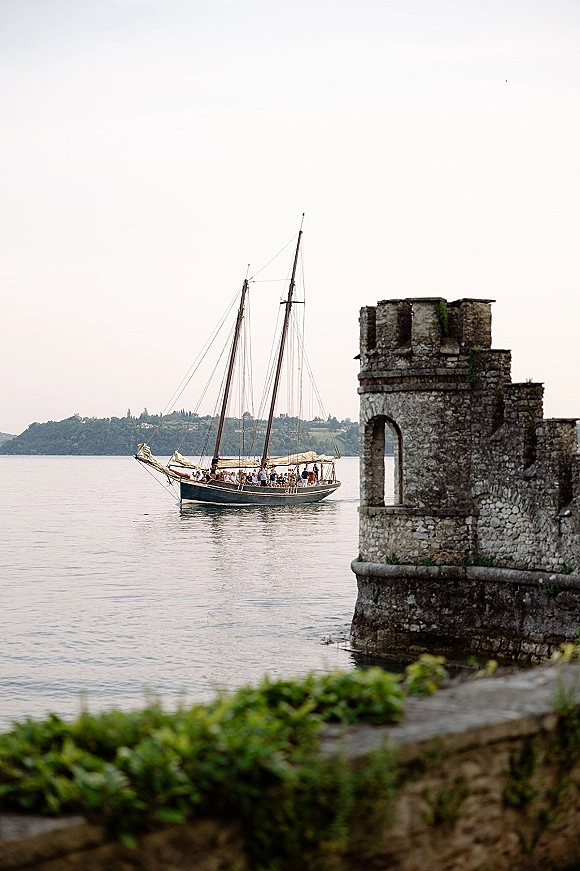 Wedding boat exit as the couple boards a wedding getaway boat sailboat, with masts and rigging on a lake by a stone castle wall