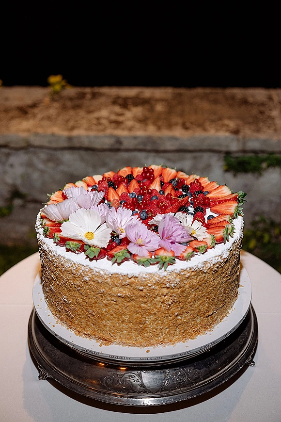 Wedding cake with fresh berry wedding cake topping, strawberries and edible flowers on a stand against a stone wall and greenery backdrop