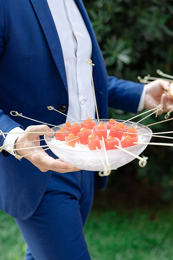 Cocktail hour appetizers in a glass bowl of crushed ice, red gelatin cubes on cocktail picks held by a caterer in a navy suit outdoors