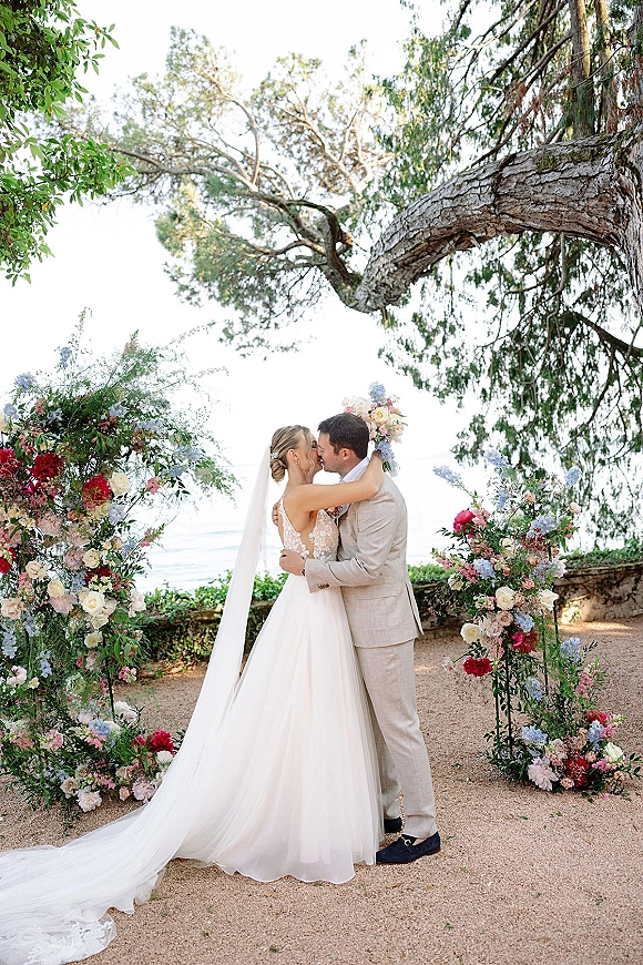 Wedding kiss portrait of bride and groom kissing beneath floral ceremony pillars, veil and lace gown flowing on a garden path with ocean view
