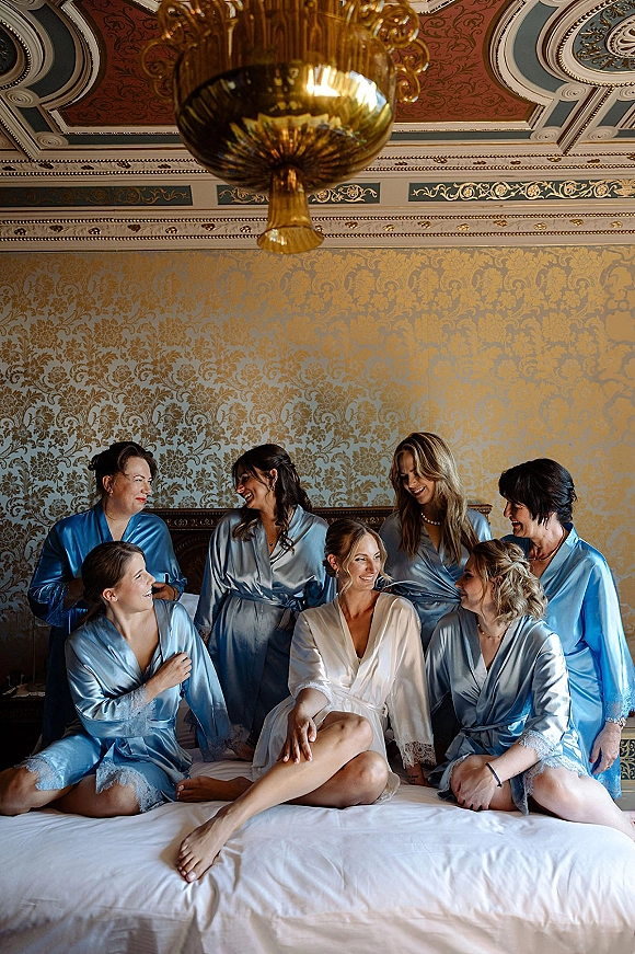 Bridesmaids getting ready in bridal party robes on a bed, laughing in satin lace-trim robes beneath an ornate chandelier in a suite