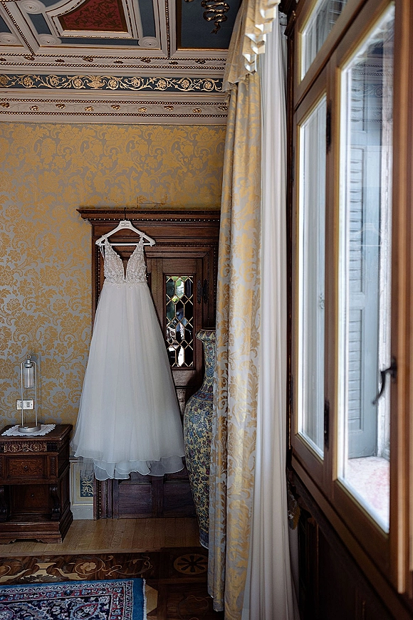 Wedding dress with lace bodice hanging from a wooden armoire, tulle skirt flowing in a vintage room with ornate wallpaper and tall windows